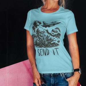 Puede incluir: Camiseta azul claro con un gr&aacute;fico negro de un soldado calavera, aviones militares y el texto "SEND IT". La camiseta es de cuello redondo.