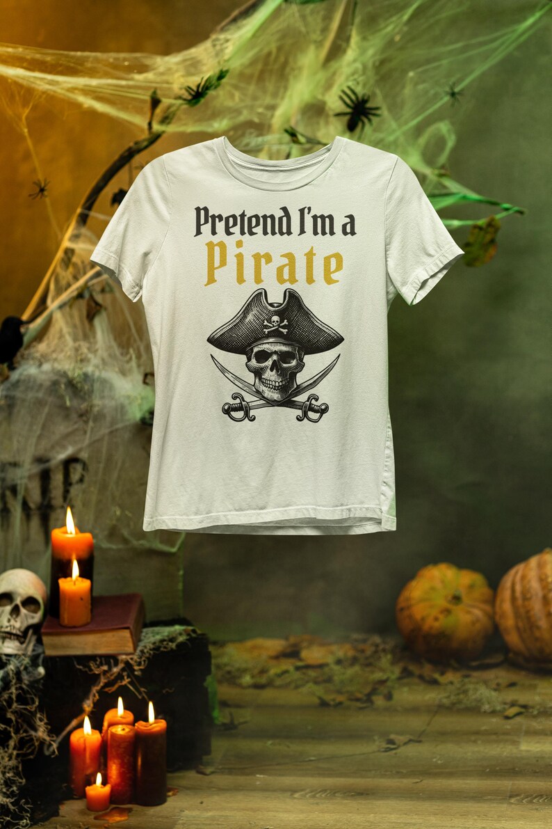 May include: White t-shirt with the text "Pretend I'm a Pirate" in black and gold. The shirt features a skull wearing a pirate hat with crossed swords. The design is centered on the front of the shirt.