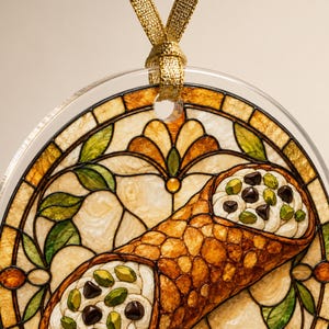 May include: A round, clear acrylic ornament featuring a stained-glass style illustration of a cannoli filled with cream, chocolate chips, and pistachios. The background displays a decorative floral pattern in amber, gold, and green tones with a gold ribbon.