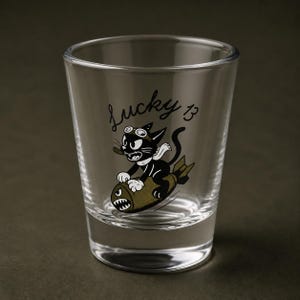 May include: A clear glass shot glass featuring a black and white cartoon cat wearing goggles and a scarf, riding an olive green bomb. The text Lucky 13 is printed in black script above the cat. The design includes a small cigar in the cat's mouth.