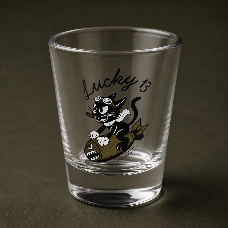May include: A clear glass shot glass featuring a black and white cartoon cat wearing goggles and a scarf, riding an olive green bomb. The text Lucky 13 is printed in black script above the cat. The design includes a small cigar in the cat's mouth.