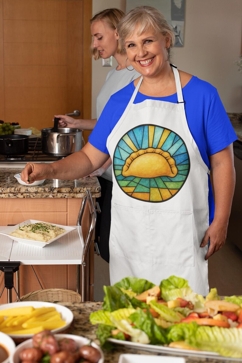May include: White apron with a colorful graphic of a golden empanada, surrounded by a sunburst design. The apron has a pocket and is worn by a person in a blue shirt, preparing food in a kitchen setting.