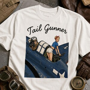May include: A white t-shirt featuring a graphic illustration of a blue military aircraft with a pilot in the cockpit and a person in a blue shirt and white shorts operating a machine gun. The text Tail Gunner is printed in black above the graphic.