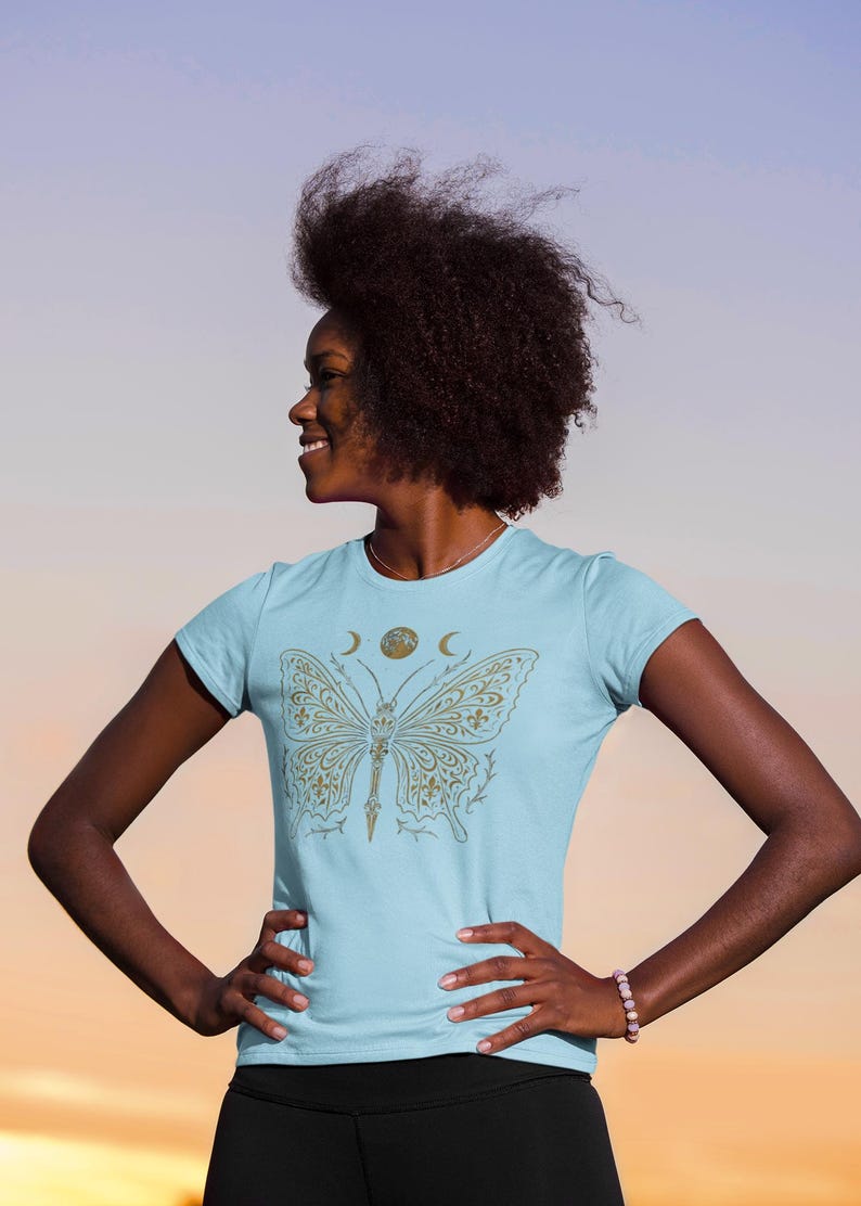 May include: Light blue t-shirt featuring a gold butterfly design. The butterfly has intricate patterns and is centered on the shirt. Above the butterfly are a crescent moon and a circle. The shirt is a crew neck and short-sleeved.