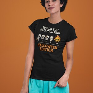 May include: Black t-shirt with the text "HOW DO YOU RATE YOUR PAIN" and "HALLOWEEN EDITION". The shirt features a pain scale with skull graphics. The person is wearing turquoise scrubs.