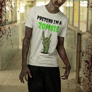 May include: White t-shirt with the text "PRETEND I'M A ZOMBIE" in black and green. The shirt features a green hand making a rock and roll gesture. The person is wearing black pinstripe pants. The background is a hallway with blood splatter.