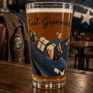 May include: A clear glass pint glass filled with amber liquid, featuring a printed graphic of a blue military aircraft with a pilot and a person in a blue shirt and white shorts operating a machine gun, with the text Tail Gunner in black script.