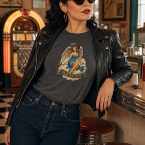 May include: A charcoal gray t-shirt featuring a graphic print of an angel with wings sitting on clouds, wearing a blue dress with a halo above her head. A banner below the illustration displays the text HEAVENLY BODY in bold capital letters.
