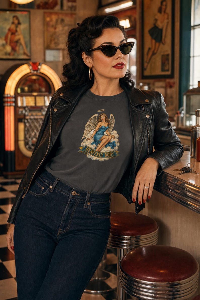 May include: A charcoal gray t-shirt featuring a graphic print of an angel with wings sitting on clouds, wearing a blue dress with a halo above her head. A banner below the illustration displays the text HEAVENLY BODY in bold capital letters.