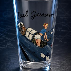 May include: A clear glass pint glass featuring a printed graphic of a blue military aircraft with a pilot in the cockpit and a figure in a blue shirt and white shorts operating a machine gun. The text Tail Gunner is printed in black script.