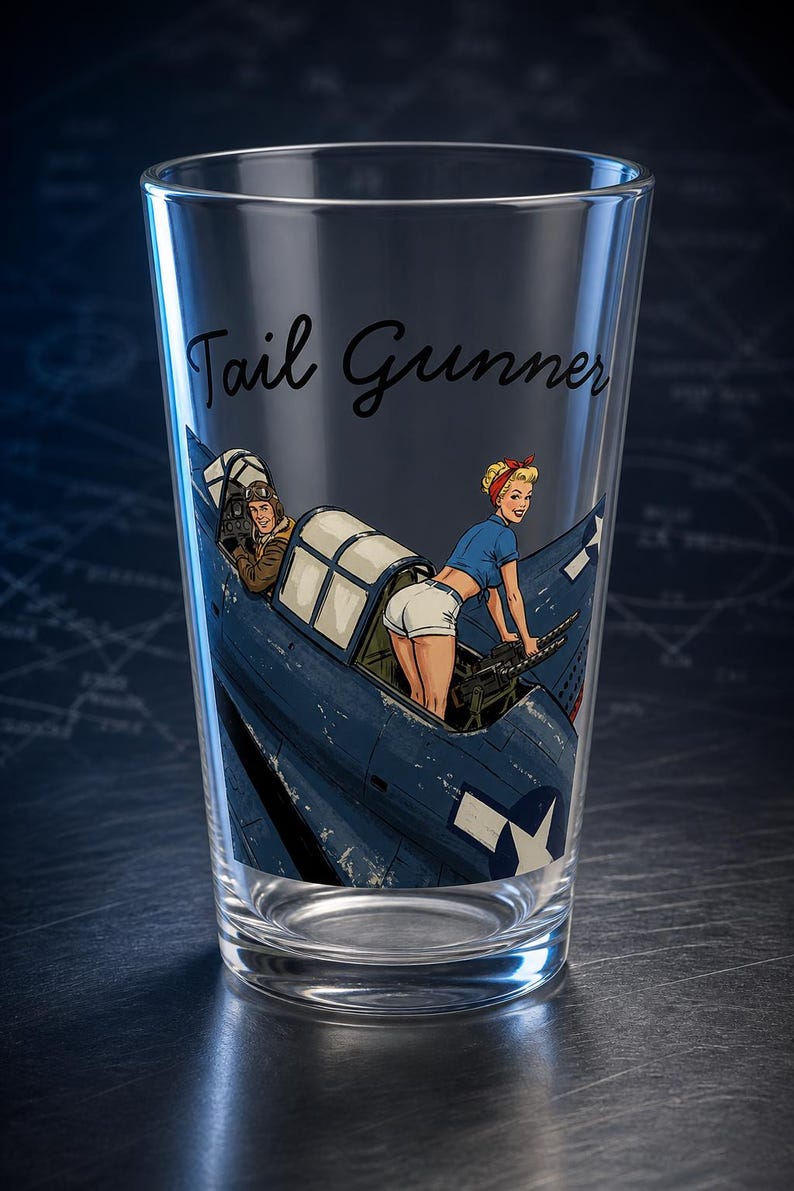 May include: A clear glass pint glass featuring a printed graphic of a blue military aircraft with a pilot in the cockpit and a figure in a blue shirt and white shorts operating a machine gun. The text Tail Gunner is printed in black script.