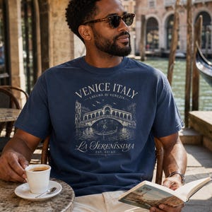 May include: A navy blue short-sleeve t-shirt featuring a cream-colored graphic print of the Rialto Bridge in Venice, Italy. The text reads VENICE ITALY, LAGUNA DI VENEZIA, LA SERENISSIMA, and EST. 421 AD, depicting a gondola and birds.