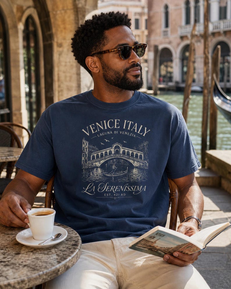 May include: A navy blue short-sleeve t-shirt featuring a cream-colored graphic print of the Rialto Bridge in Venice, Italy. The text reads VENICE ITALY, LAGUNA DI VENEZIA, LA SERENISSIMA, and EST. 421 AD, depicting a gondola and birds.