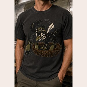 Puede incluir: Una camiseta de manga corta gris carb&oacute;n con una ilustraci&oacute;n gr&aacute;fica de un p&aacute;jaro negro con gafas de aviador en un nido. El p&aacute;jaro lleva una bufanda blanca y est&aacute; rodeado por tres objetos de color verde oliva con forma de bomba.