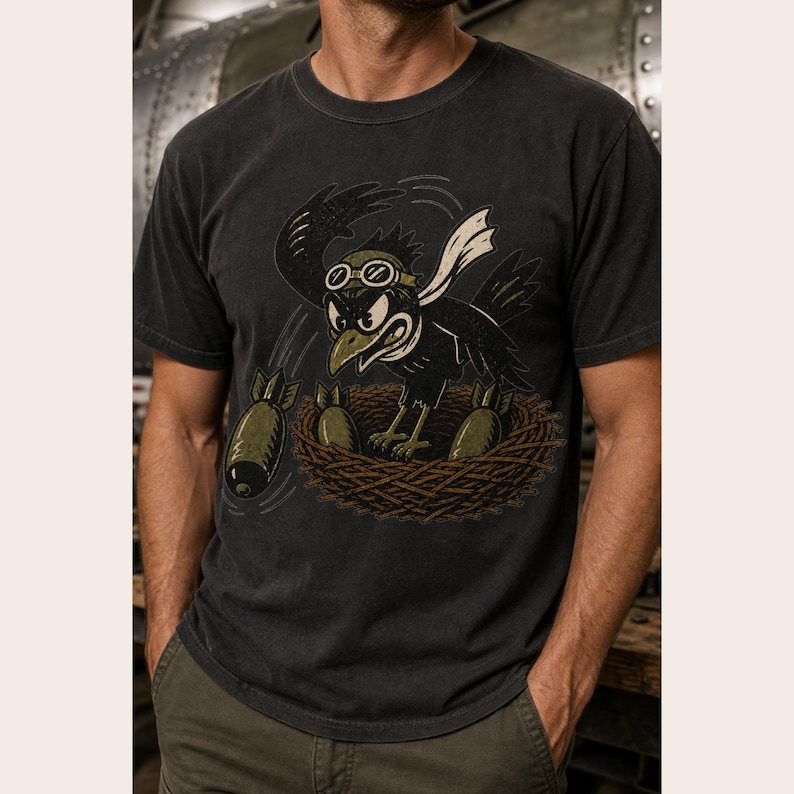 Puede incluir: Una camiseta de manga corta gris carb&oacute;n con una ilustraci&oacute;n gr&aacute;fica de un p&aacute;jaro negro con gafas de aviador en un nido. El p&aacute;jaro lleva una bufanda blanca y est&aacute; rodeado por tres objetos de color verde oliva con forma de bomba.