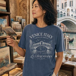 May include: A blue short-sleeve t-shirt featuring a white graphic print of the Rialto Bridge in Venice, Italy. The text reads "VENICE ITALY", "LAGUNA DI VENEZIA", "LA SERENISSIMA", and "EST. 421 AD" with an illustration of a gondola and birds.