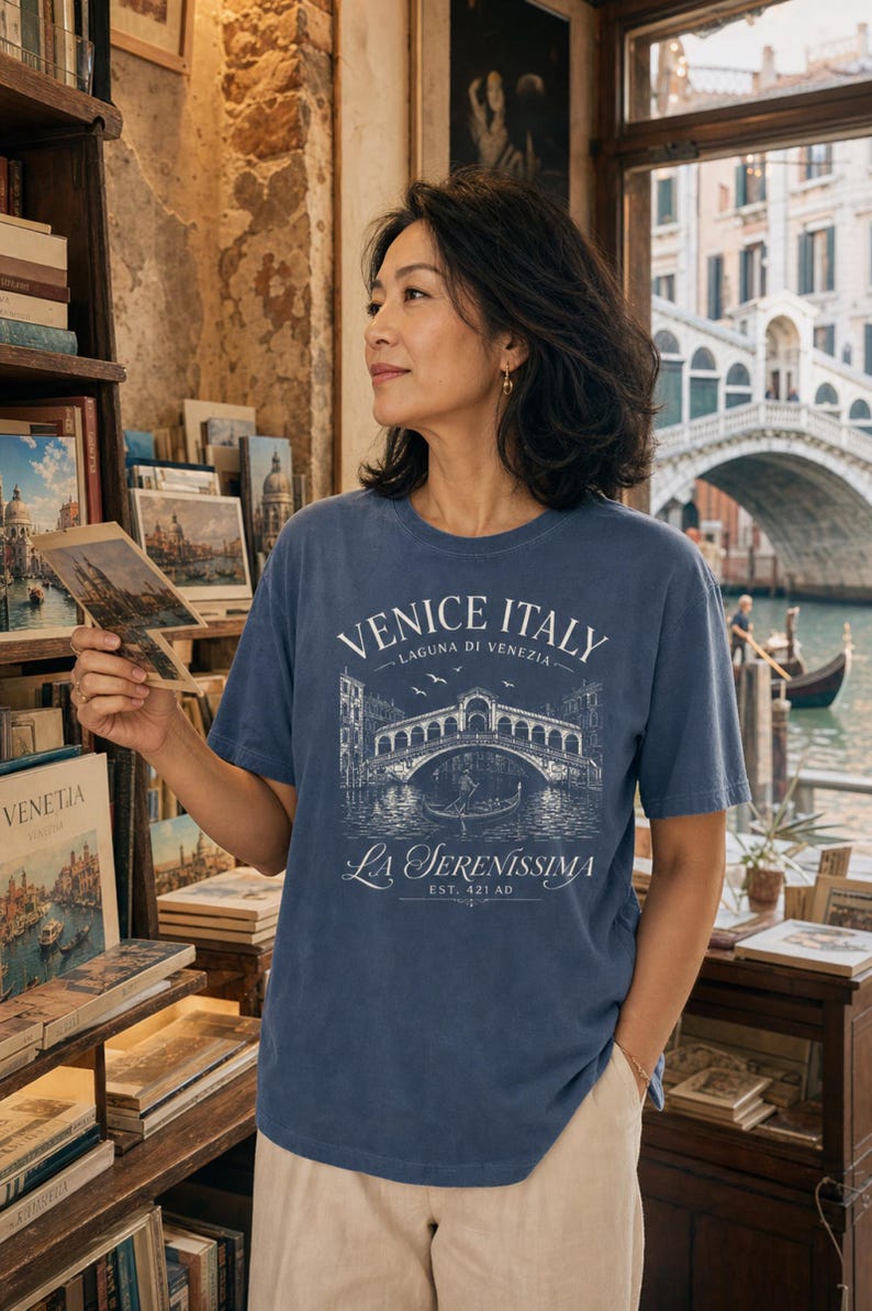 May include: A blue short-sleeve t-shirt featuring a white graphic print of the Rialto Bridge in Venice, Italy. The text reads "VENICE ITALY", "LAGUNA DI VENEZIA", "LA SERENISSIMA", and "EST. 421 AD" with an illustration of a gondola and birds.