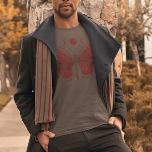 May include: A gray t-shirt featuring a red butterfly design with moon and sun details. The shirt is worn with a black coat, scarf, and pants. The person is leaning against a stone wall.