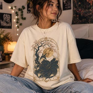 May include: An off-white short-sleeve t-shirt featuring a graphic print of a multi-tailed fox sitting under a large moon and a flowering tree. The illustration is framed by an ornate archway design in shades of dark blue, gold, and cream.