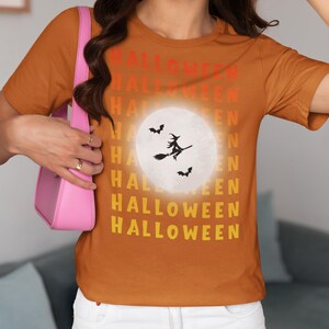 Puede incluir: Camiseta naranja con la palabra "HALLOWEEN" apilada verticalmente en rojo, naranja y amarillo. Un gr&aacute;fico de luna blanca presenta una bruja en una escoba y murci&eacute;lagos. La camiseta es de cuello redondo y manga corta.