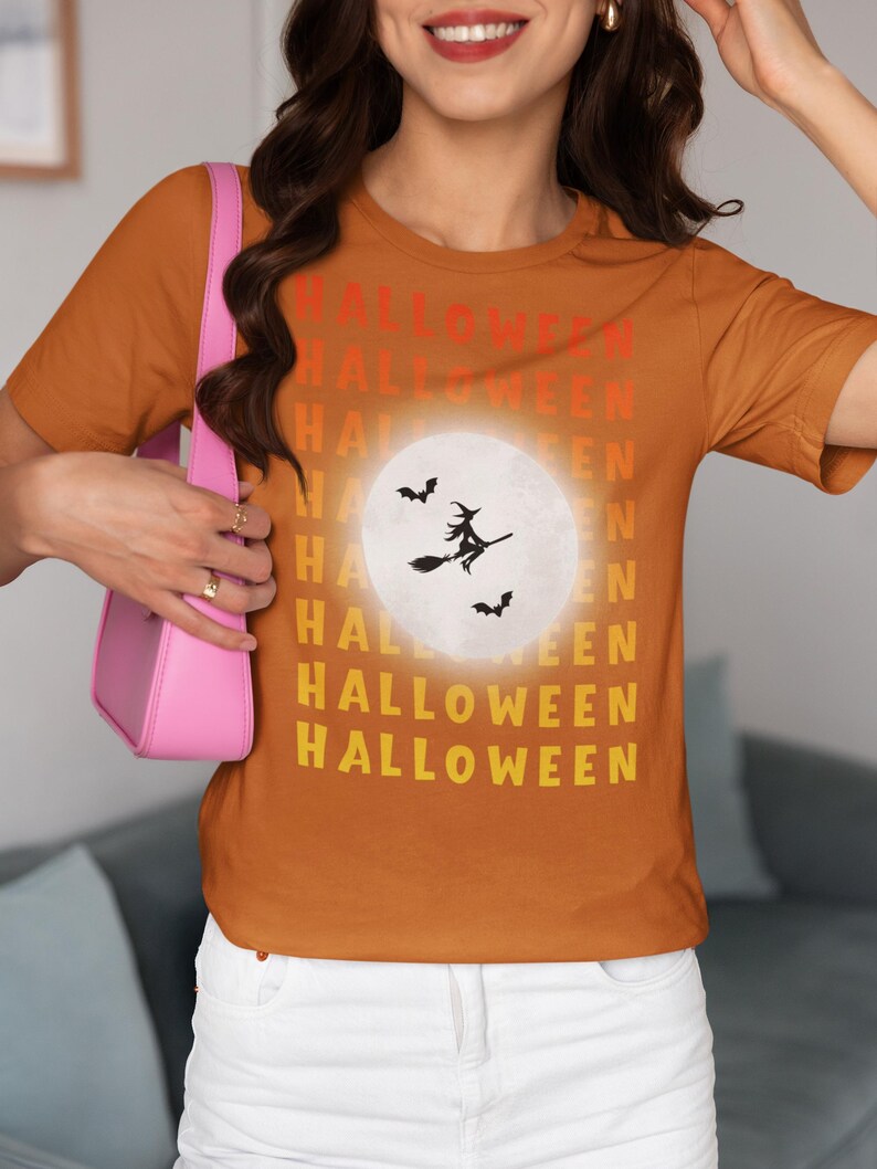 Puede incluir: Camiseta naranja con la palabra "HALLOWEEN" apilada verticalmente en rojo, naranja y amarillo. Un gr&aacute;fico de luna blanca presenta una bruja en una escoba y murci&eacute;lagos. La camiseta es de cuello redondo y manga corta.
