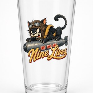 Op de afbeelding: Een helder glazen drinkglas met een afbeelding van een zwarte kat met een bruine vliegeniersmuts en een bril, rijdend op een grijze bom. Onder de bom staat de tekst Nine Lives in een geel en rood lettertype met drie kleine zonnesymbolen.