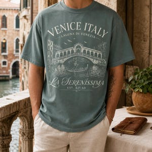 May include: A dusty blue short-sleeve t-shirt featuring a white graphic print of the Rialto Bridge in Venice, Italy. The text reads VENICE ITALY, LAGUNA DI VENEZIA, LA SERENISSIMA, and EST. 421 AD, depicting a gondola and birds in the scene.