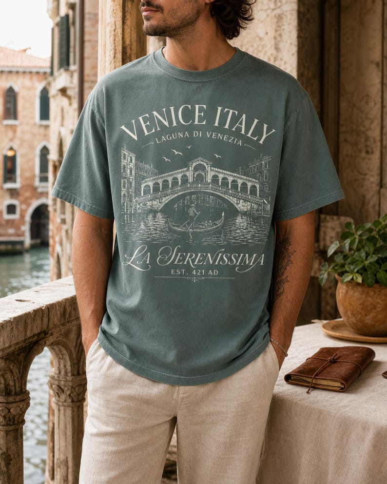 May include: A dusty blue short-sleeve t-shirt featuring a white graphic print of the Rialto Bridge in Venice, Italy. The text reads VENICE ITALY, LAGUNA DI VENEZIA, LA SERENISSIMA, and EST. 421 AD, depicting a gondola and birds in the scene.