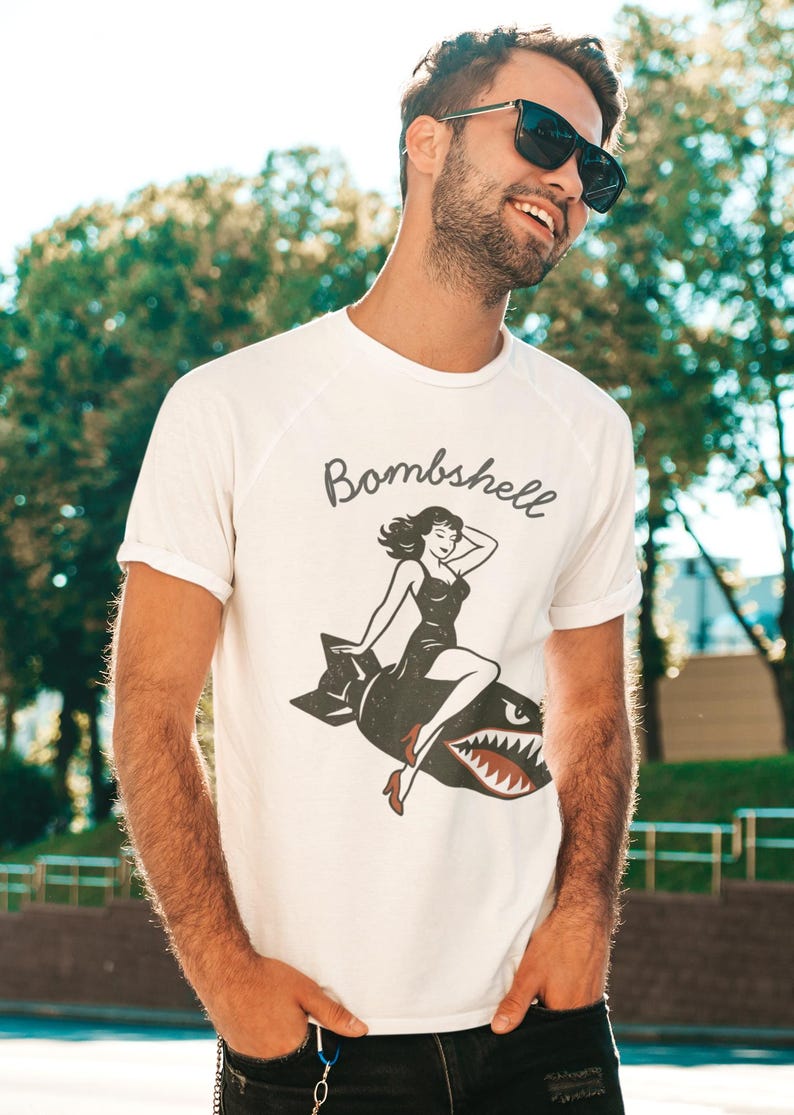 May include: White t-shirt with a vintage-style graphic of a woman riding a bomb, with the word "Bombshell" above the image. The design features a woman in a black dress and red heels. The shirt is a classic crew neck.