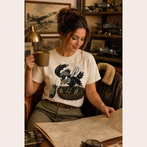 Puede incluir: Una camiseta de manga corta de color crema con una ilustraci&oacute;n gr&aacute;fica de un p&aacute;jaro negro con gafas de aviador y una bufanda blanca, sentado en un nido mientras suelta bombas verdes. El dise&ntilde;o est&aacute; centrado en la parte del pecho.