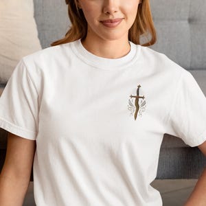 May include: White t-shirt with a detailed illustration of a sword with a cross-shaped hilt and decorative elements. The design is in shades of brown and gold. The shirt is made of a soft, comfortable material.