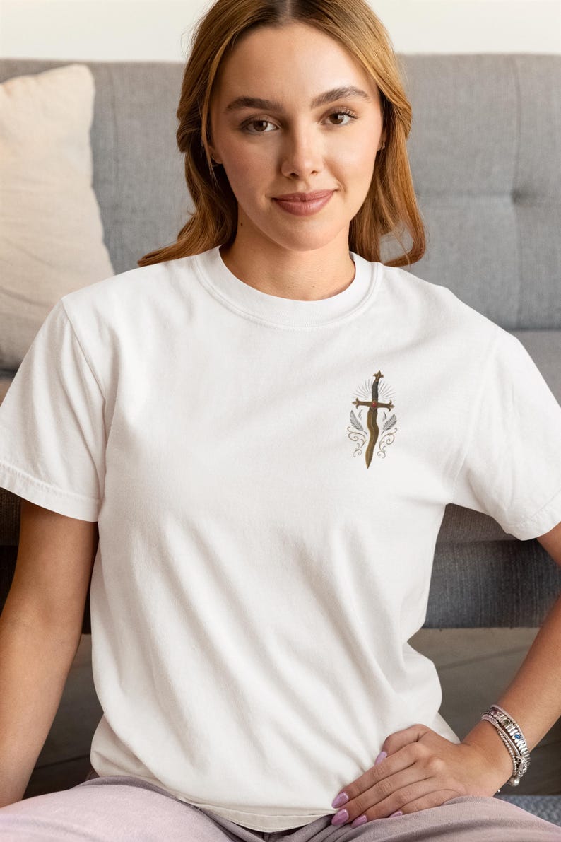 May include: White t-shirt with a detailed illustration of a sword with a cross-shaped hilt and decorative elements. The design is in shades of brown and gold. The shirt is made of a soft, comfortable material.