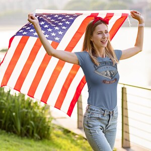 Puede incluir: Una camiseta de manga corta color azul pizarra con un estampado gr&aacute;fico de un bombardero furtivo B-2 Spirit y el texto America The Beautiful y 250 Years. La camiseta se combina con vaqueros azul claro y una cinta roja para el pelo.