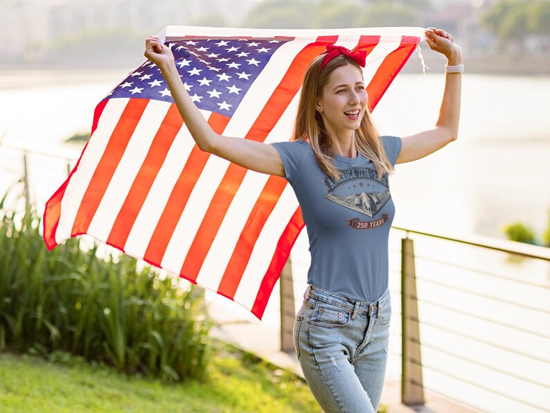 Puede incluir: Una camiseta de manga corta color azul pizarra con un estampado gr&aacute;fico de un bombardero furtivo B-2 Spirit y el texto America The Beautiful y 250 Years. La camiseta se combina con vaqueros azul claro y una cinta roja para el pelo.