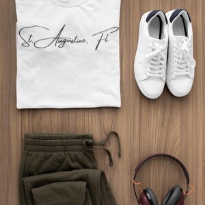 Peut inclure: Vue de dessus de v&ecirc;tements et accessoires d&eacute;contract&eacute;s sur une surface en bois. Comprend un t-shirt blanc avec "St. Augustine, FL" en &eacute;criture manuscrite, un pantalon de surv&ecirc;tement vert olive, des baskets blanches et un casque marron.
