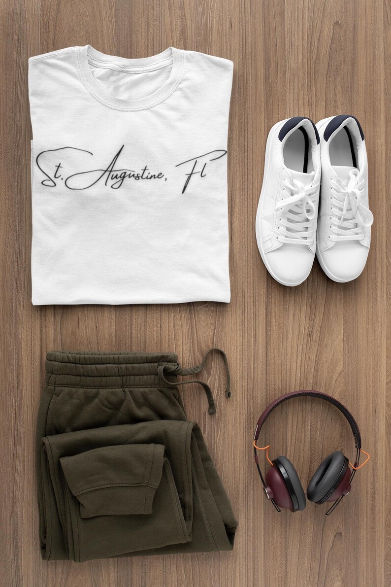 Peut inclure: Vue de dessus de v&ecirc;tements et accessoires d&eacute;contract&eacute;s sur une surface en bois. Comprend un t-shirt blanc avec "St. Augustine, FL" en &eacute;criture manuscrite, un pantalon de surv&ecirc;tement vert olive, des baskets blanches et un casque marron.