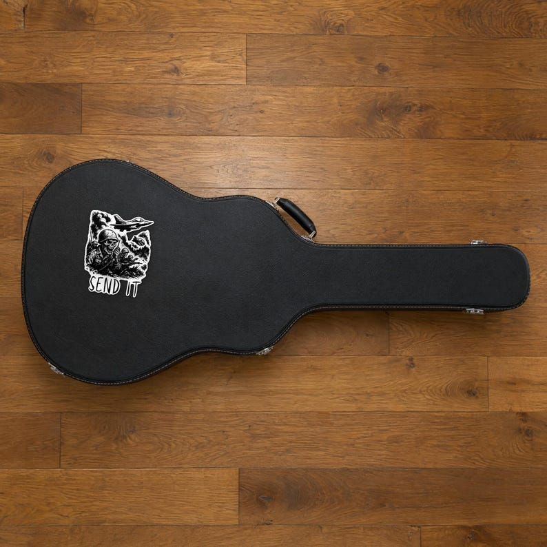Może przedstawiać: Czarny futerał na gitarę z białą naklejką przedstawiającą grafikę czaszki w hełmie wojskowym, samolot myśliwski oraz napis SEND IT wydrukowany pogrubioną wielką czcionką w dolnej części naklejki.