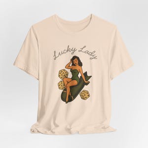 May include: A light beige t-shirt featuring a graphic illustration of a person in a green swimsuit sitting on a dark bomb, surrounded by three dice. The text "Lucky Lady" is printed in a dark cursive font above the central illustration.