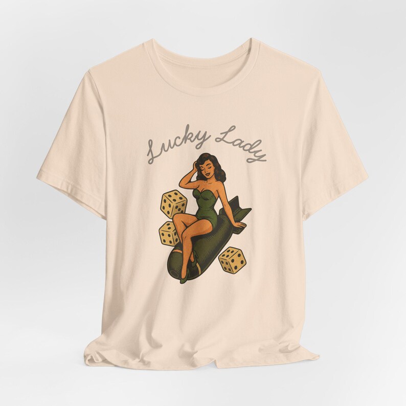 May include: A light beige t-shirt featuring a graphic illustration of a person in a green swimsuit sitting on a dark bomb, surrounded by three dice. The text "Lucky Lady" is printed in a dark cursive font above the central illustration.