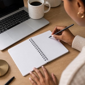 May include: A white spiral-bound notebook with lined pages lies open on a wooden desk. A black pen is held in position to write on the paper. Nearby, a silver laptop, a speckled white ceramic mug filled with black coffee, and a black pen are visible.