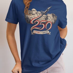 May include: A navy blue short-sleeve t-shirt featuring a graphic of Mount Rushmore with a jet flying above. The design includes the number 250 in red and white, with a banner below displaying the years 1776-2026 in a vintage-style font.