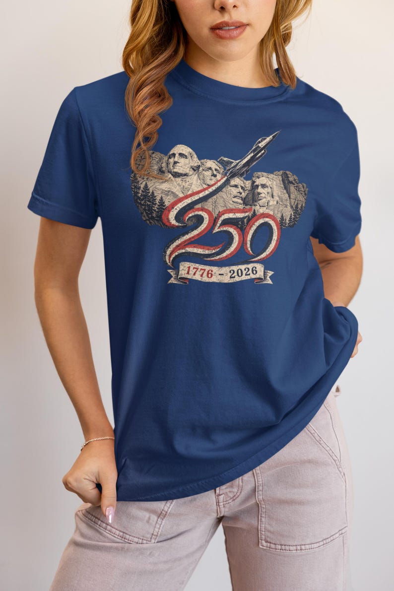 May include: A navy blue short-sleeve t-shirt featuring a graphic of Mount Rushmore with a jet flying above. The design includes the number 250 in red and white, with a banner below displaying the years 1776-2026 in a vintage-style font.