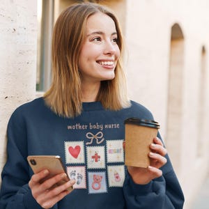 K&ouml;nnte beinhalten: Marineblaues Sweatshirt mit dem Text "mother baby nurse" und mehreren Briefmarken-Grafiken. Die Grafiken umfassen ein Herz, Fu&szlig;abdr&uuml;cke, einen Teddyb&auml;ren und eine Babyflasche. Ein Einweg-Kaffeebecher wird in der Hand gehalten.