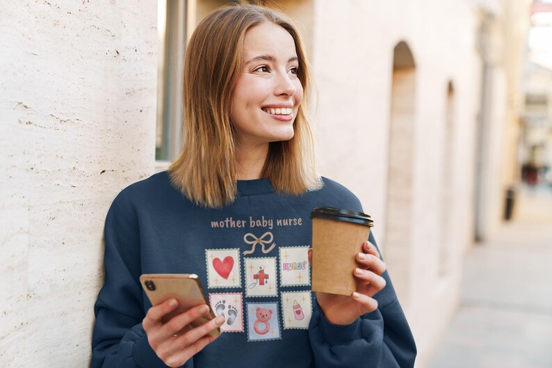 K&ouml;nnte beinhalten: Marineblaues Sweatshirt mit dem Text "mother baby nurse" und mehreren Briefmarken-Grafiken. Die Grafiken umfassen ein Herz, Fu&szlig;abdr&uuml;cke, einen Teddyb&auml;ren und eine Babyflasche. Ein Einweg-Kaffeebecher wird in der Hand gehalten.