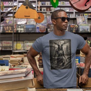 May include: A slate blue short-sleeve t-shirt featuring a rectangular graphic print of a dark, skeletal, antlered creature standing in a forest under a full moon. The design is rendered in shades of black, gray, and white with high contrast details.