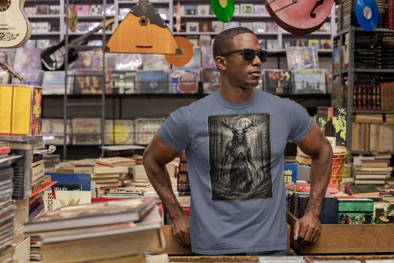 May include: A slate blue short-sleeve t-shirt featuring a rectangular graphic print of a dark, skeletal, antlered creature standing in a forest under a full moon. The design is rendered in shades of black, gray, and white with high contrast details.