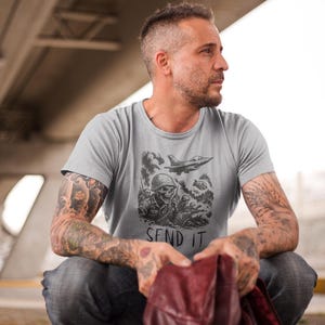 Puede incluir: Camiseta gris claro con un gr&aacute;fico de tem&aacute;tica militar. El dise&ntilde;o presenta un soldado calavera, un avi&oacute;n a reacci&oacute;n y el texto "SEND IT" en blanco y negro. La camiseta est&aacute; hecha de un material suave.
