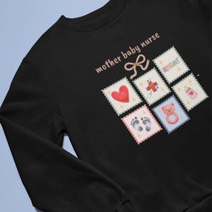K&ouml;nnte beinhalten: Schwarzes Sweatshirt mit dem Text "mother baby nurse" und einer Schleifengrafik. Das Design umfasst Briefmarken-&auml;hnliche Bilder eines Herzens, Fu&szlig;abdr&uuml;cke, eines Teddyb&auml;ren, einer Babyflasche, eines Kreuzes und das Wort "nurse".