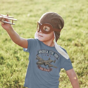 May include: A blue t-shirt featuring a graphic of a vintage military airplane with the text America 1776-2026 and 250 Years. The shirt is worn with a brown aviator cap and goggles, while a small wooden toy airplane is held in the air.