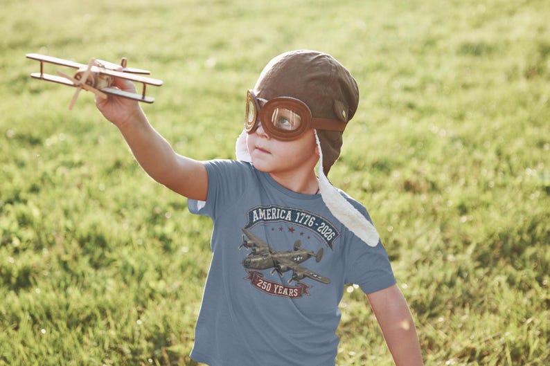 May include: A blue t-shirt featuring a graphic of a vintage military airplane with the text America 1776-2026 and 250 Years. The shirt is worn with a brown aviator cap and goggles, while a small wooden toy airplane is held in the air.